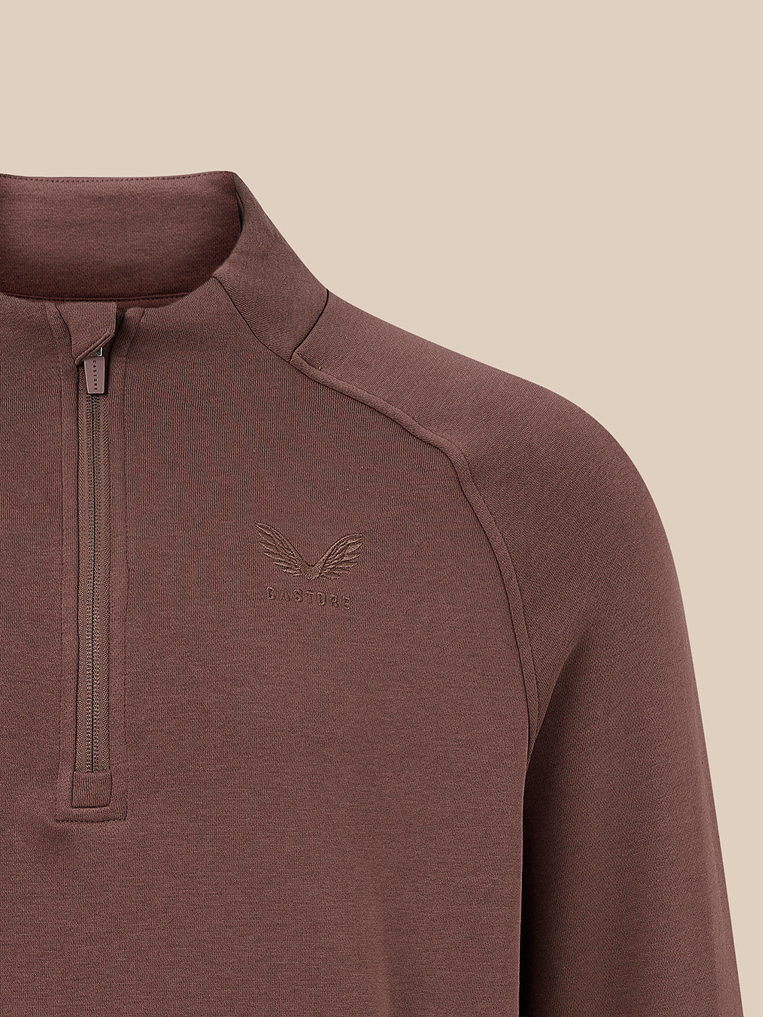Close-up of a brown quarter-zip pullover sweater with a subtle embroidered winged logo on the left chest, set against a solid beige background.