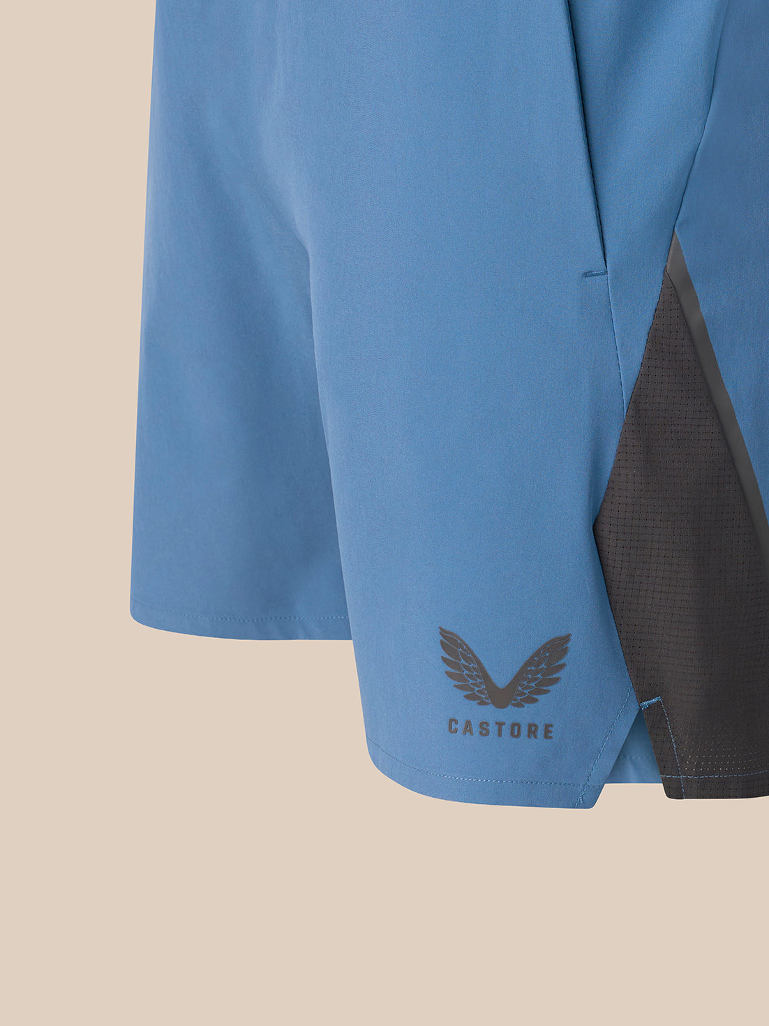 Close-up of a light blue athletic shorts leg with a dark gray mesh side panel and a dark gray Castore wing logo near the hem, set against a solid beige background.