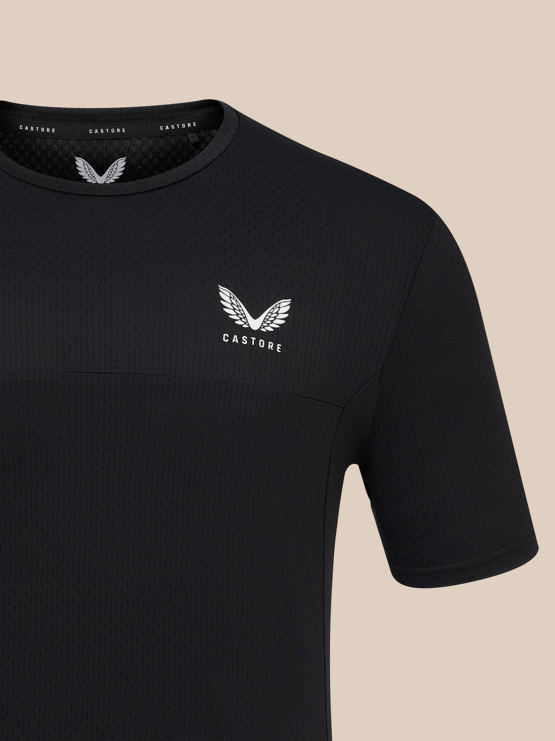 A close-up of a black short-sleeve athletic t-shirt with a textured waffle knit pattern, featuring a white winged logo and the brand name 'CASTORE' on the left chest, set against a solid beige background.