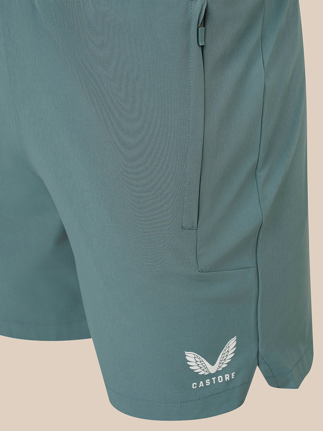 Close-up of teal athletic shorts with a side zip pocket and a white Castore wing logo near the lower hem, shown against a plain beige background.