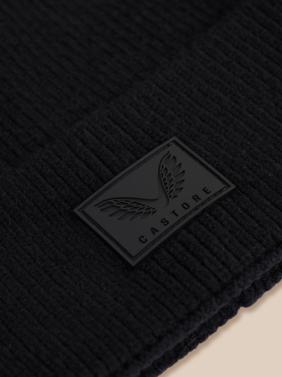 Close-up of a black ribbed knit beanie with a black rubber patch featuring a winged logo and the brand name CASTORE.