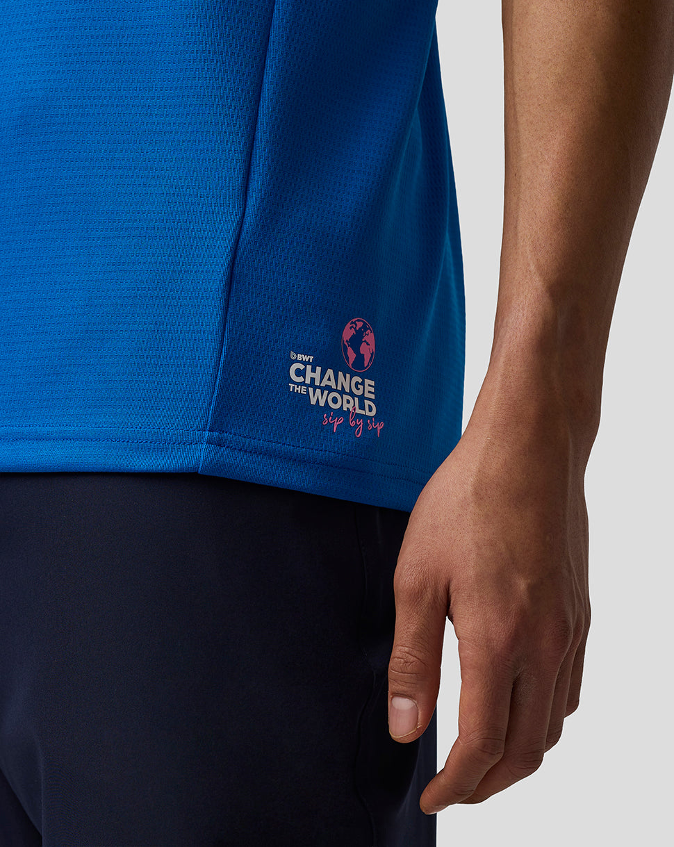 Close-up of a person wearing a bright blue athletic top and dark navy shorts, with the sleeve cuff showing a small white and pink 'Change the World' graphic and the person's hand visible against a light gray background.