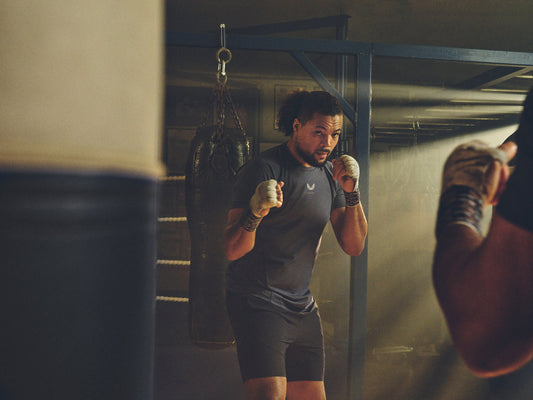 Boxer in a gym wearing a dark athletic shirt and wrapped fists, raised in fighting stance beside a hanging punching bag.