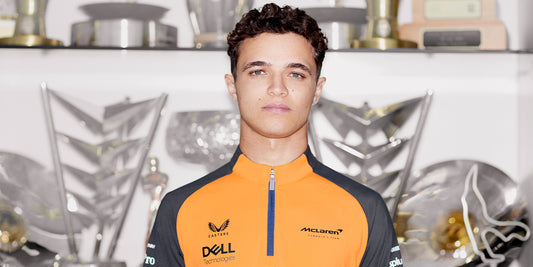 Young man in a bright orange and navy Castore McLaren jacket with trophies in the background
