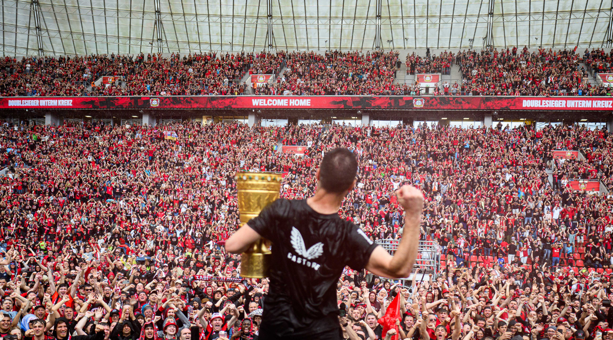 Athlete in a black Castore hoodie holds up a gold trophy before a packed stadium of red clad fans.