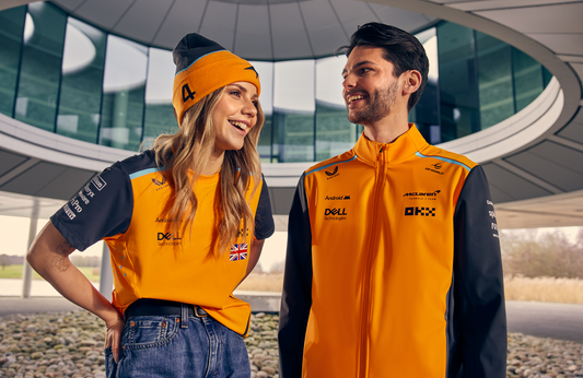 Smiling woman and man in orange McLaren teamwear jackets pose in front of a modern curved glass building.