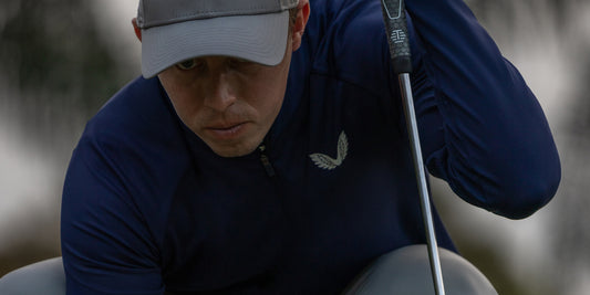 Close up of a golfer in navy Castore apparel and a gray cap, gripping a golf club.