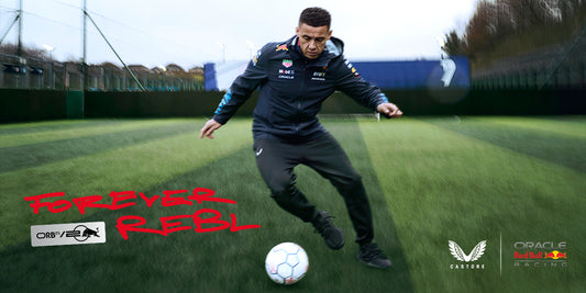 Athlete wearing Oracle Red Bull Racing x Castore gear kicks a soccer ball on a training pitch with Forever Rebel branding