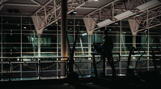 Person on a treadmill in a dimly lit gym with large windows and string lights along the railing.