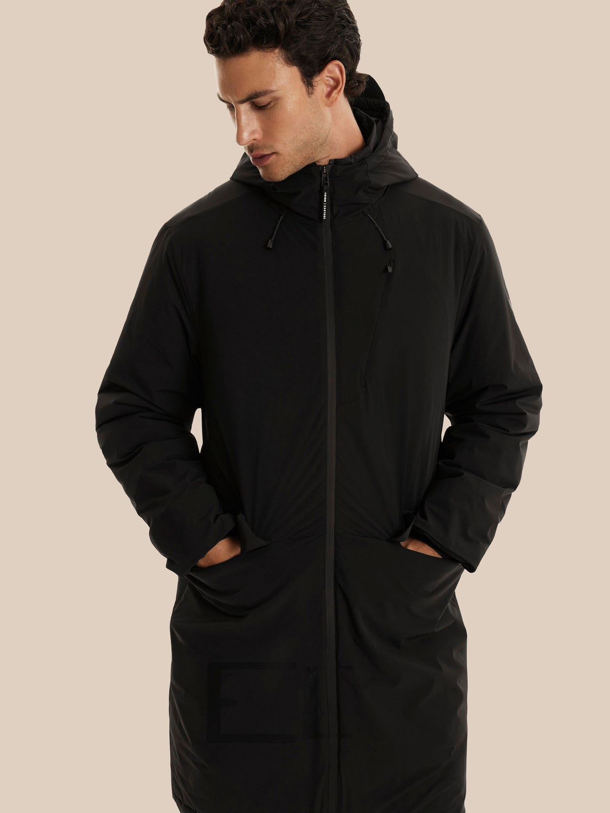 A man wearing a long black hooded puffer jacket with a full front zipper and large front pockets, posed against a solid beige background.