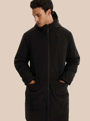 A man wearing a long black hooded puffer jacket with a full front zipper and large front pockets, posed against a solid beige background.
