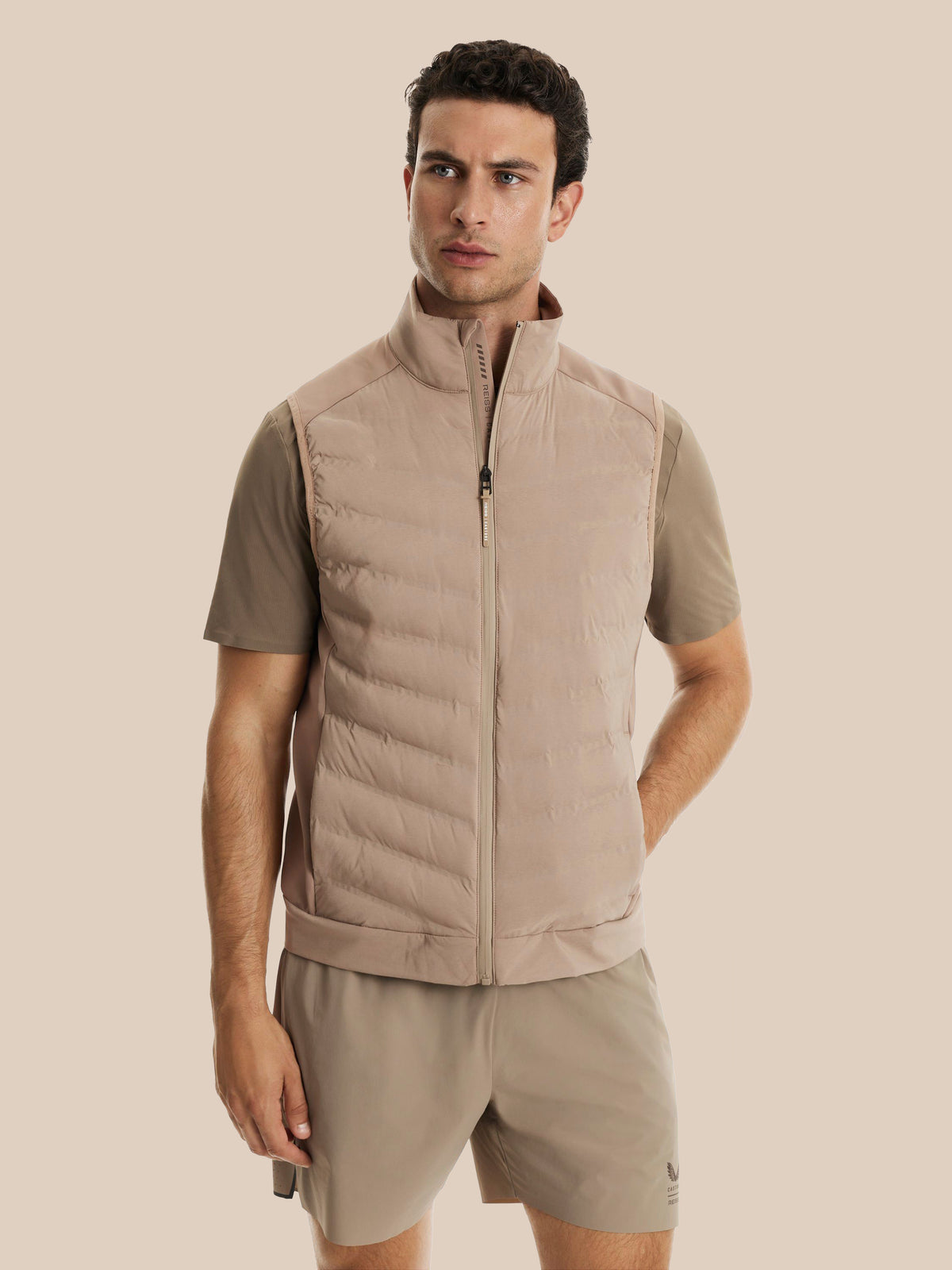 A man wearing a light beige puffer vest over a taupe short-sleeve shirt and matching beige shorts, standing against a solid beige background.