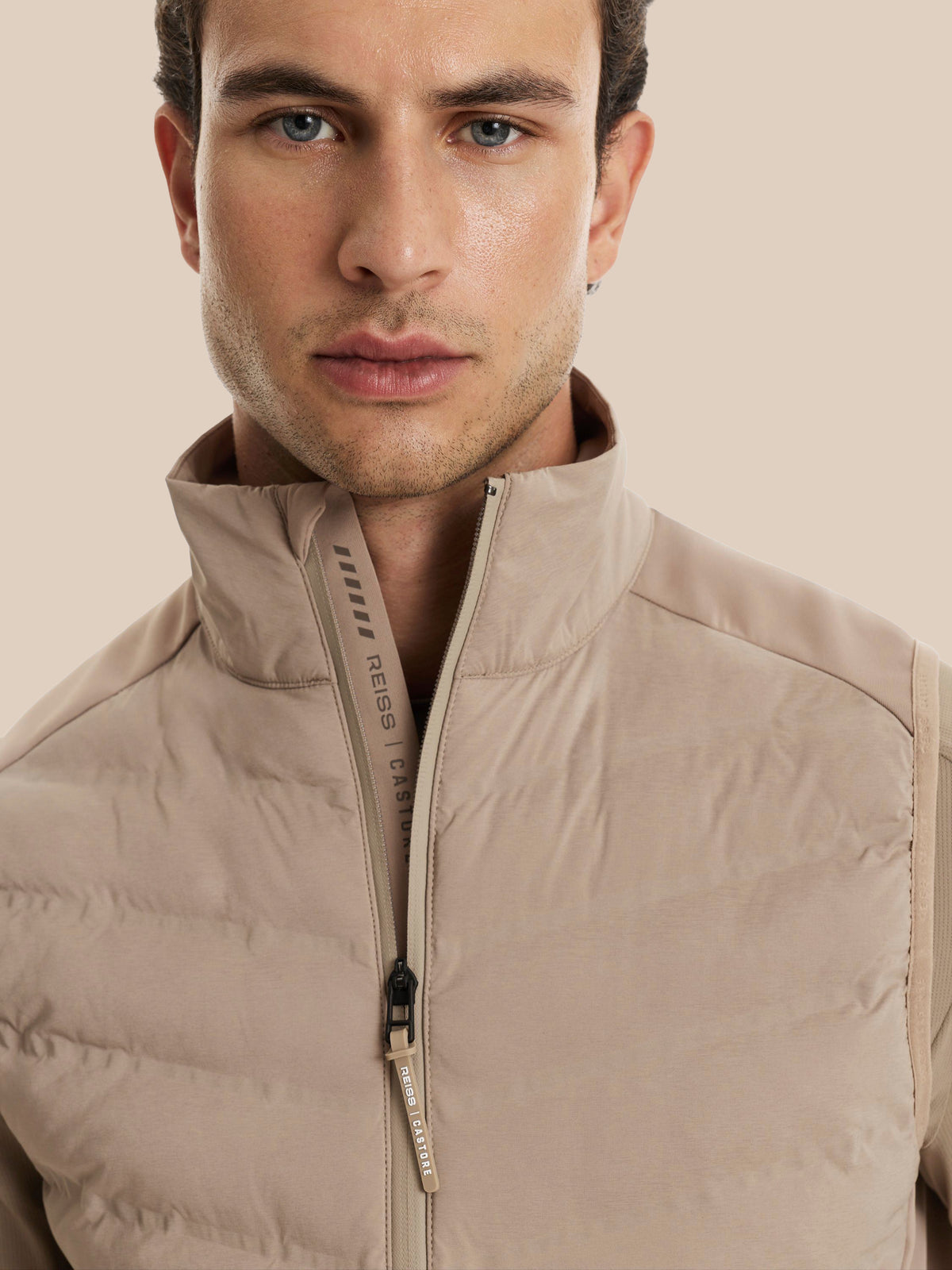 A close-up of a man wearing a beige puffer vest with a high stand-up collar and a front zipper, set against a solid beige background.