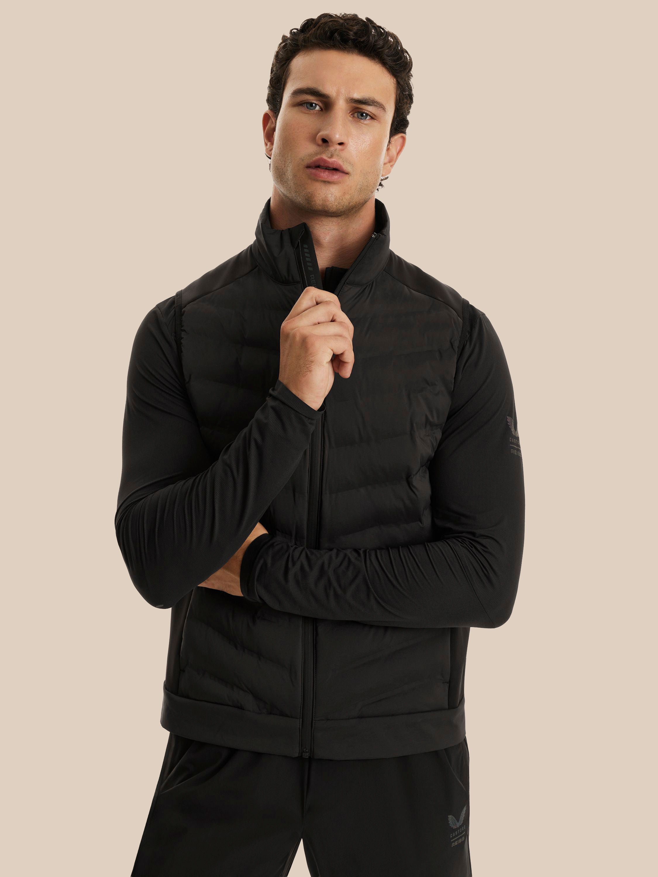 A man wearing a black long-sleeve zip-up athletic jacket with a stand collar and black pants, posed against a solid beige studio background.