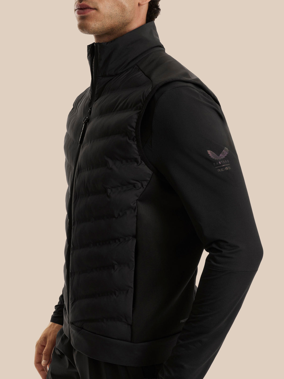 A man wearing a black zip-front puffer vest over a black long-sleeve top, shown in a side profile against a plain beige studio background. A small dark logo is visible on the upper left sleeve.