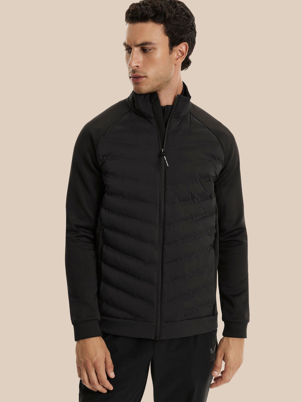 A man wearing a black zip-up puffer jacket with a stand collar and horizontal quilting, paired with black pants, set against a solid beige background.