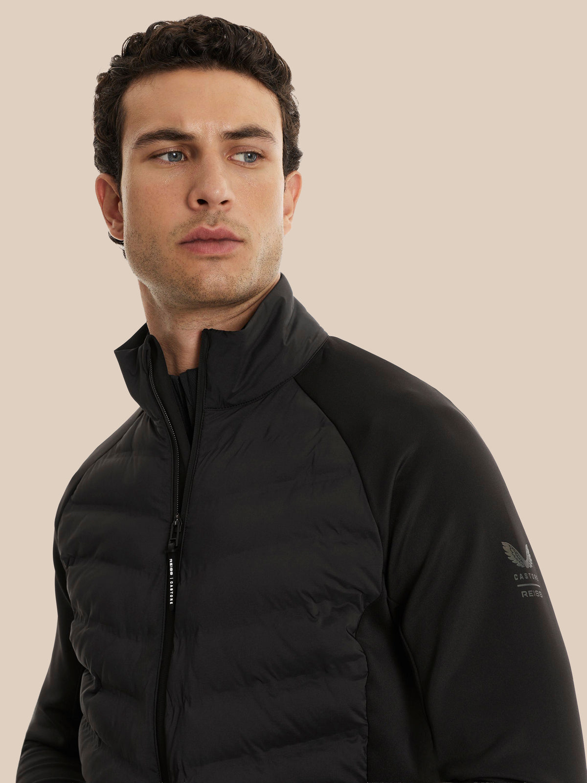 A man wearing a black CASTORE puffer jacket with a high collar and zippered front, shown from the chest up against a solid beige background.