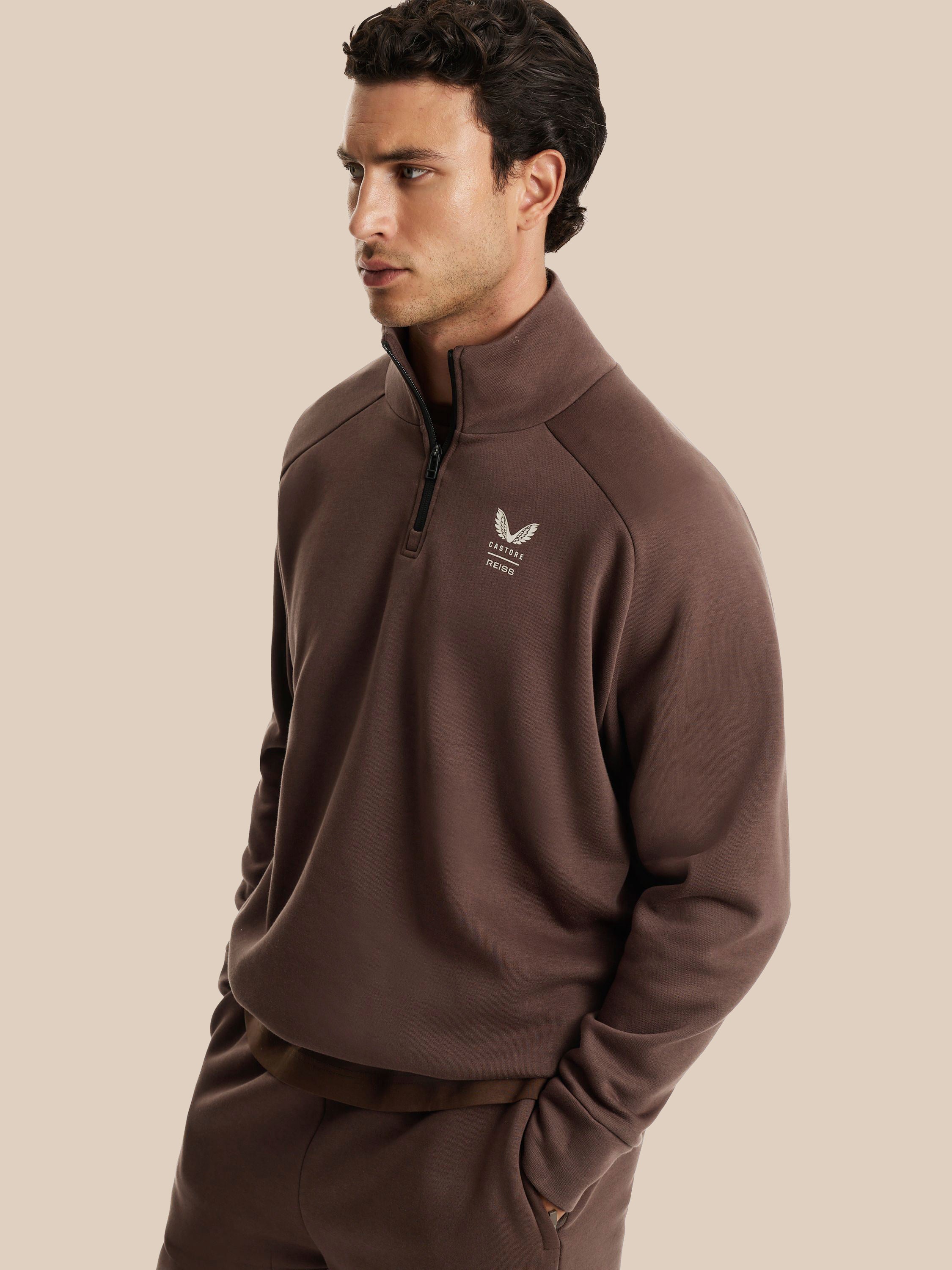 A man wearing a brown quarter-zip fleece pullover with a small white embroidered logo on the left chest, paired with matching brown pants, set against a solid beige background.