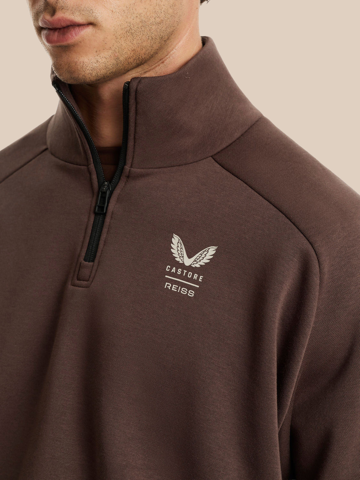 A close-up of a man wearing a brown quarter-zip pullover sweatshirt with a white embroidered winged logo and the text 'CASTORE REISS' on the left chest, set against a solid beige background.