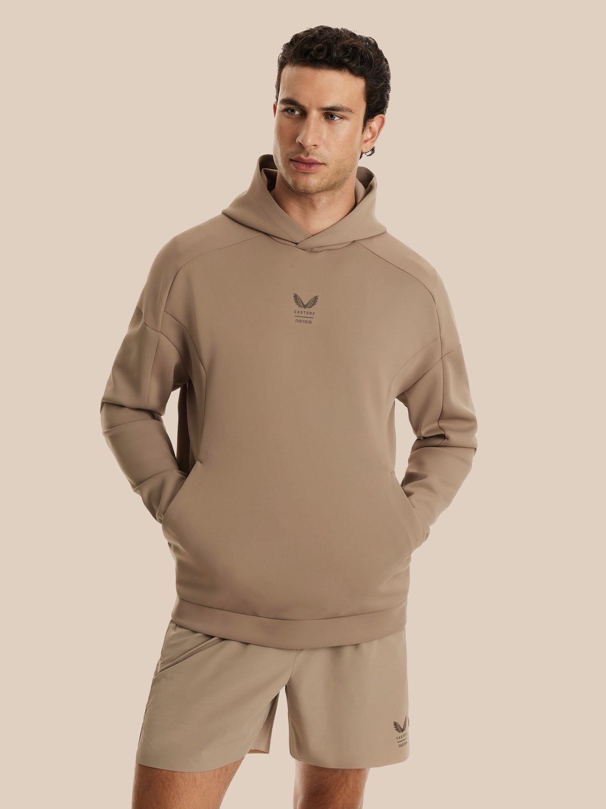 A man wearing a taupe-beige pullover hoodie with a small dark logo on the chest, paired with matching beige shorts, against a solid light beige background.
