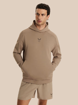 A man wearing a taupe-beige pullover hoodie with a small dark logo on the chest, paired with matching beige shorts, against a solid light beige background.