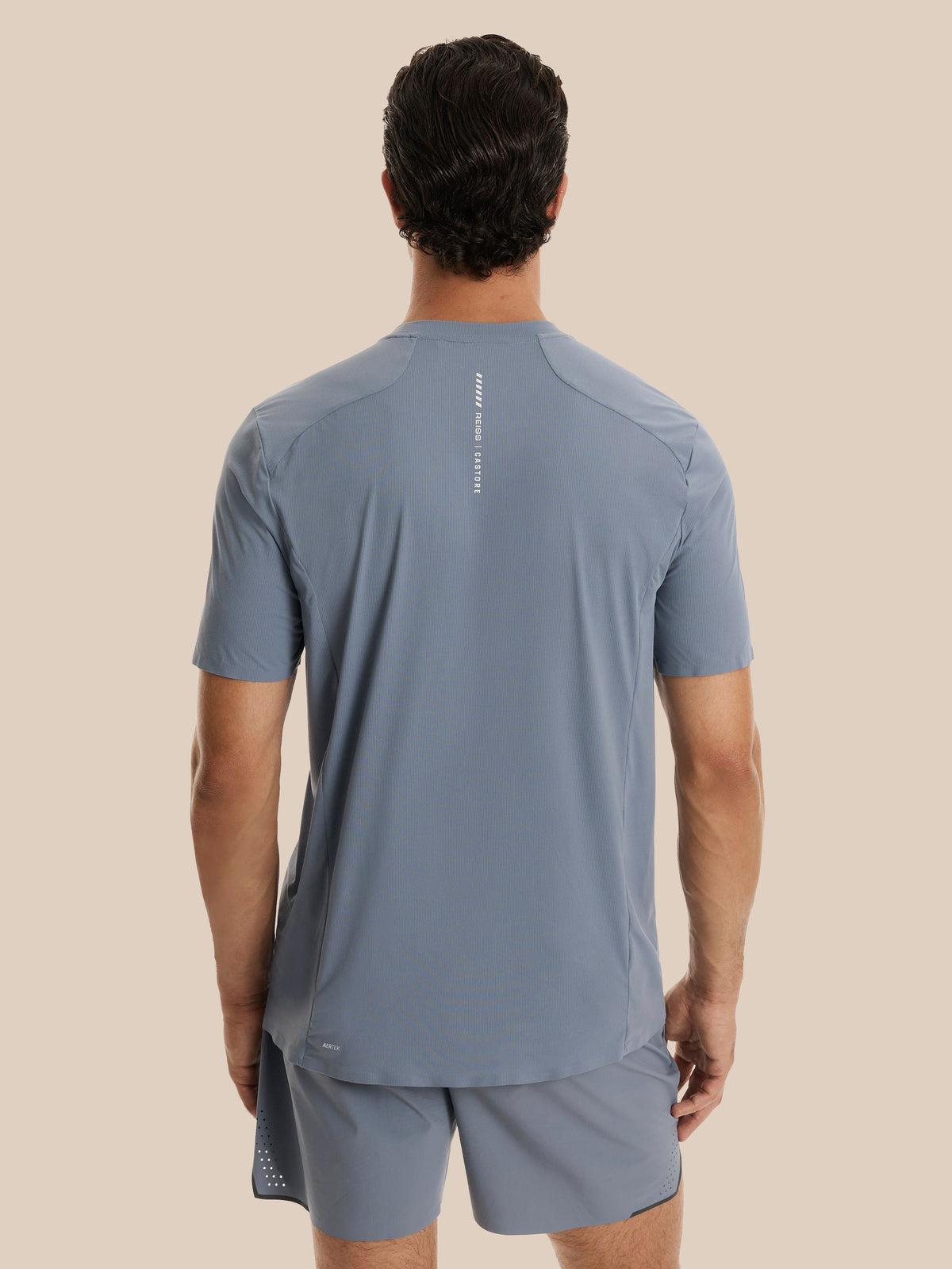A man viewed from behind wearing a slate blue athletic t-shirt and matching shorts against a solid beige background.
