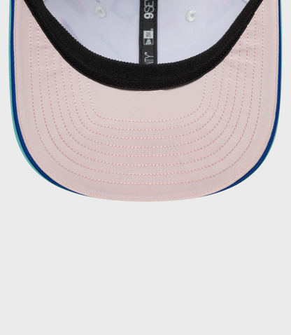 Close up of pastel pink crown with black edge and blue top button on a teal brim cap.