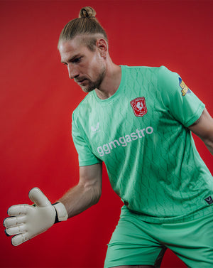 Goalkeeper wearing FC Twente 2025-2026 green jersey prepares a save against a red backdrop