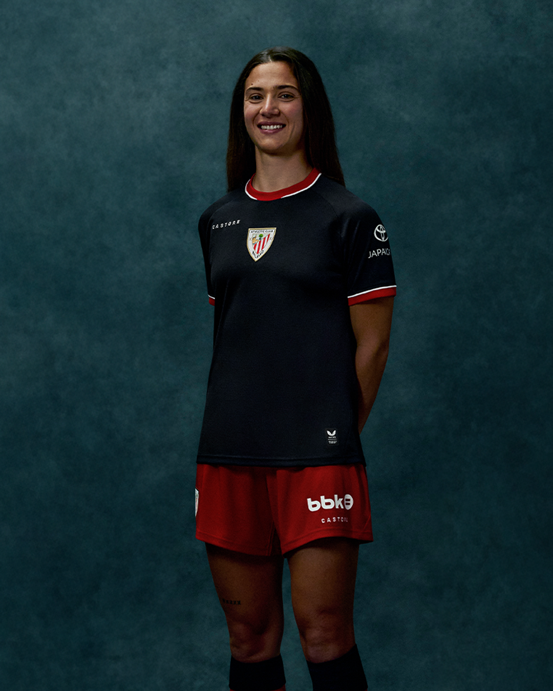 Portrait of a female athlete wearing the Athletic Club 26/27 away shirt in navy with red trim against a teal backdrop