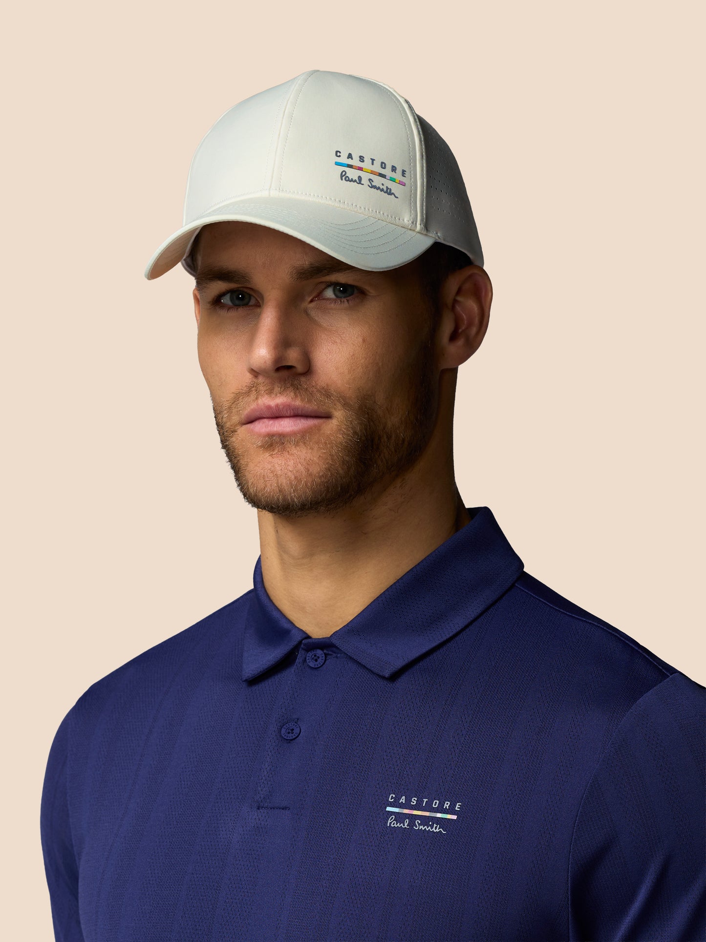 Man wearing a blue polo shirt and beige cap with brand logos on a beige background