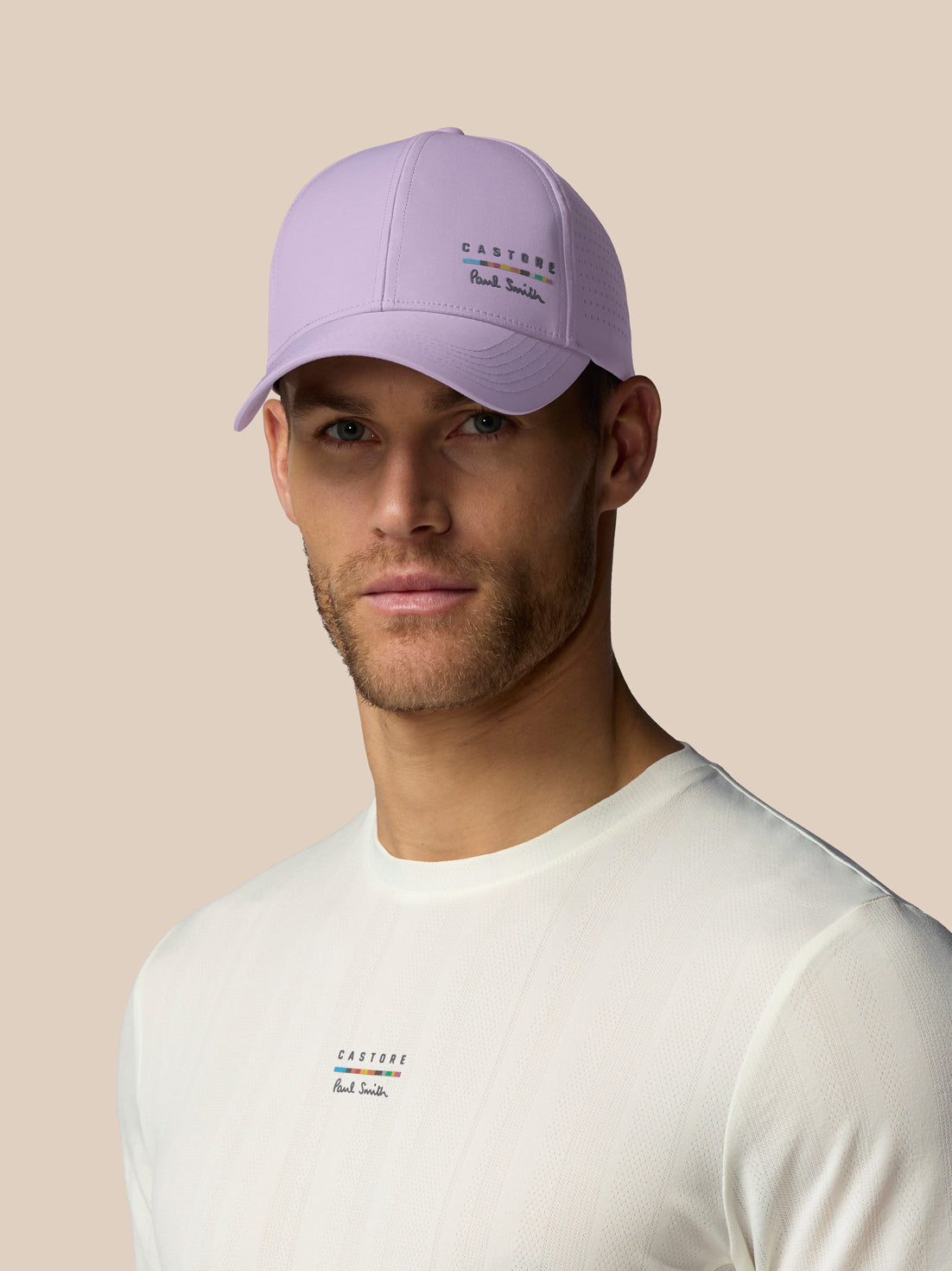Man wearing a light purple cap and white shirt with brand logos on a beige background