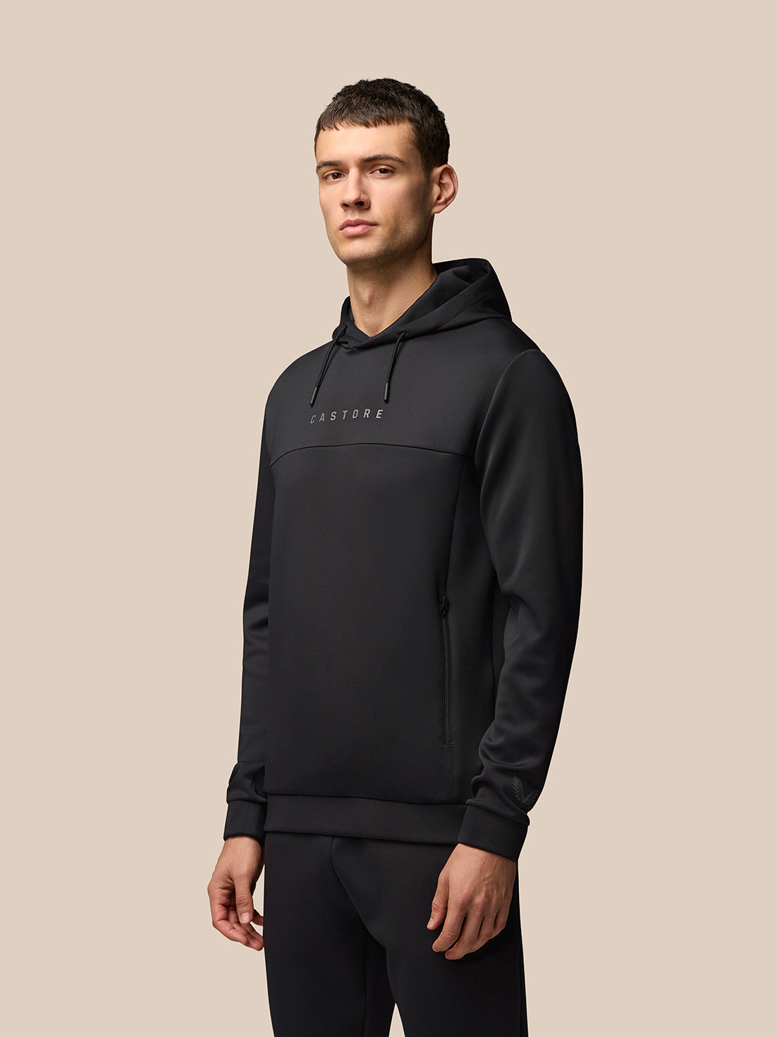 Male model in a black Castore Flex Lifestyle Hoodie with chest logo against a beige background.