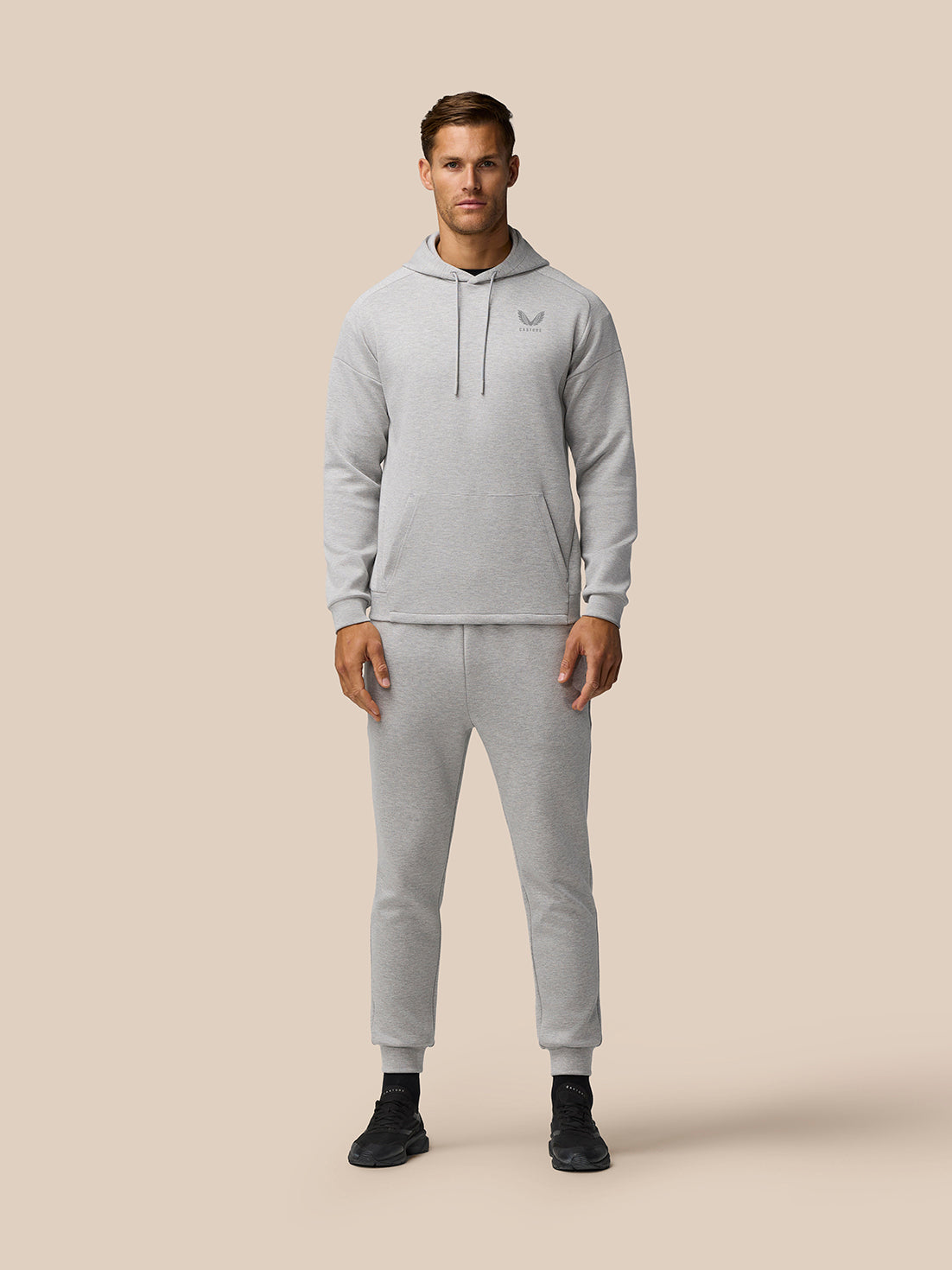 Male model wearing grey marl travel lifestyle hoodie and matching joggers, showcasing relaxed fit and cuffed hems on a beige background.