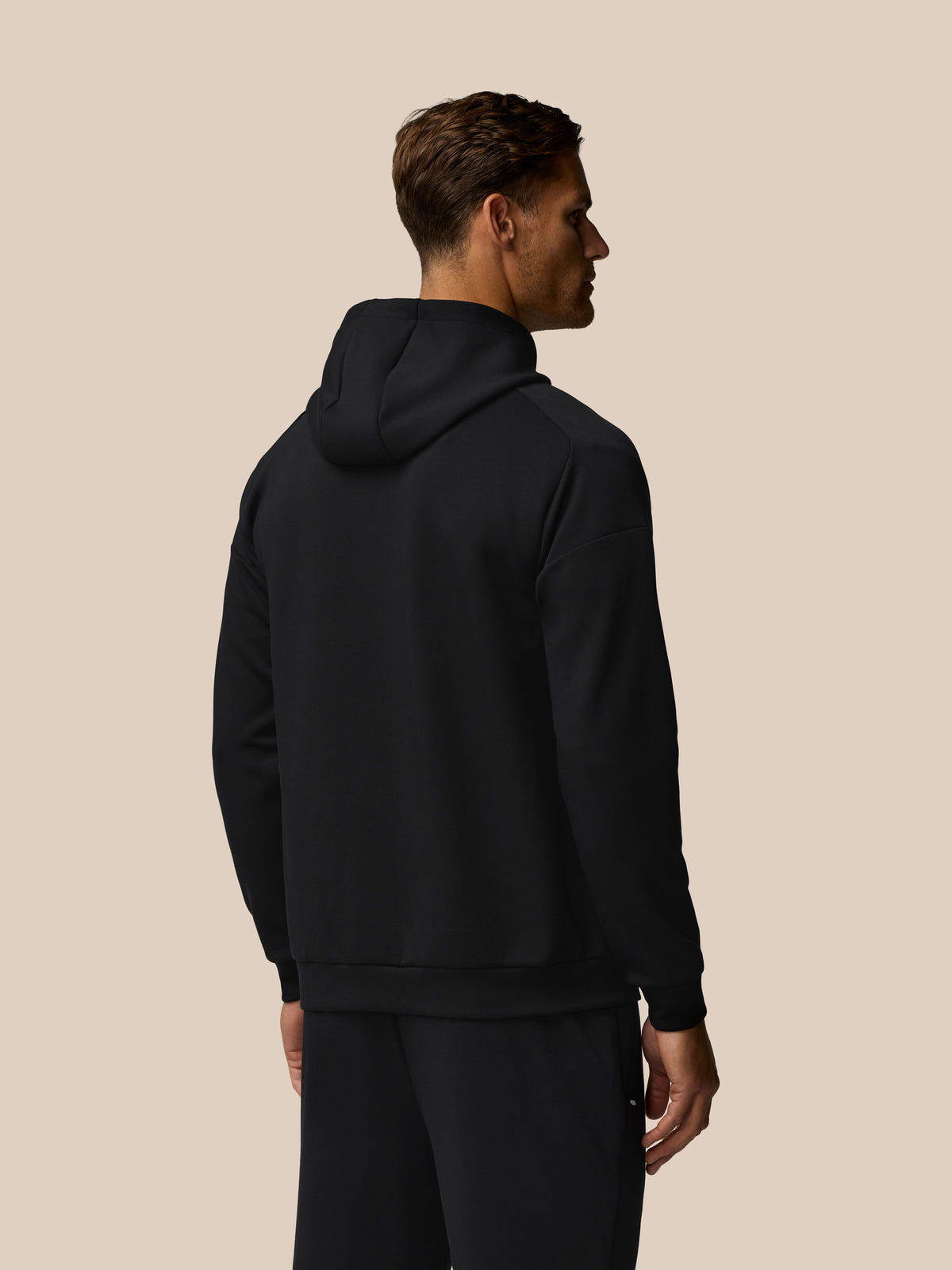 Back view of male wearing black Travel Lifestyle Hoodie with hood down against beige backdrop