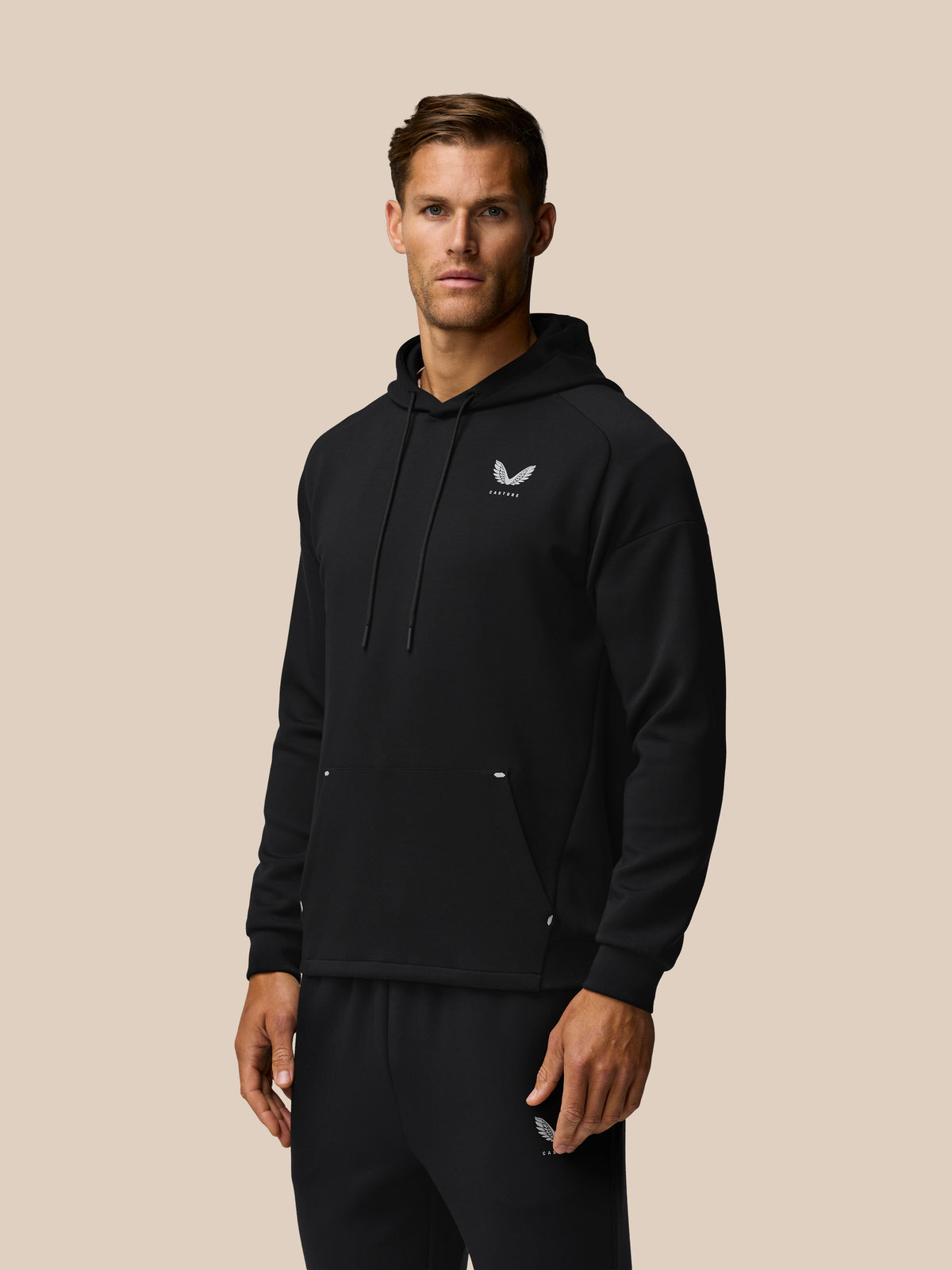 Male model in black Travel Lifestyle Hoodie shown slightly angled, chest logo and drawstring hood visible
