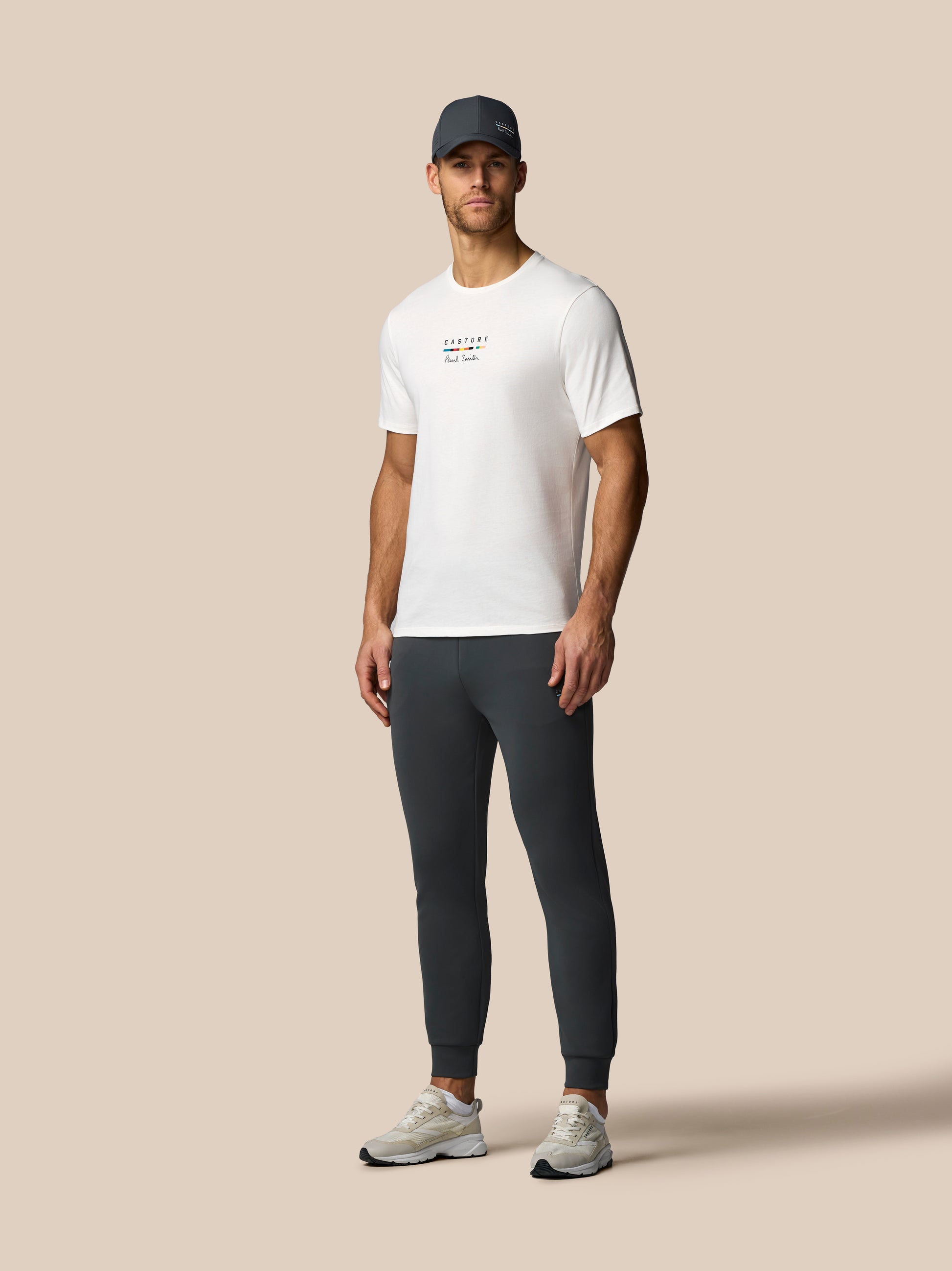 Man wearing a white t-shirt, dark pants, and a cap on a beige background