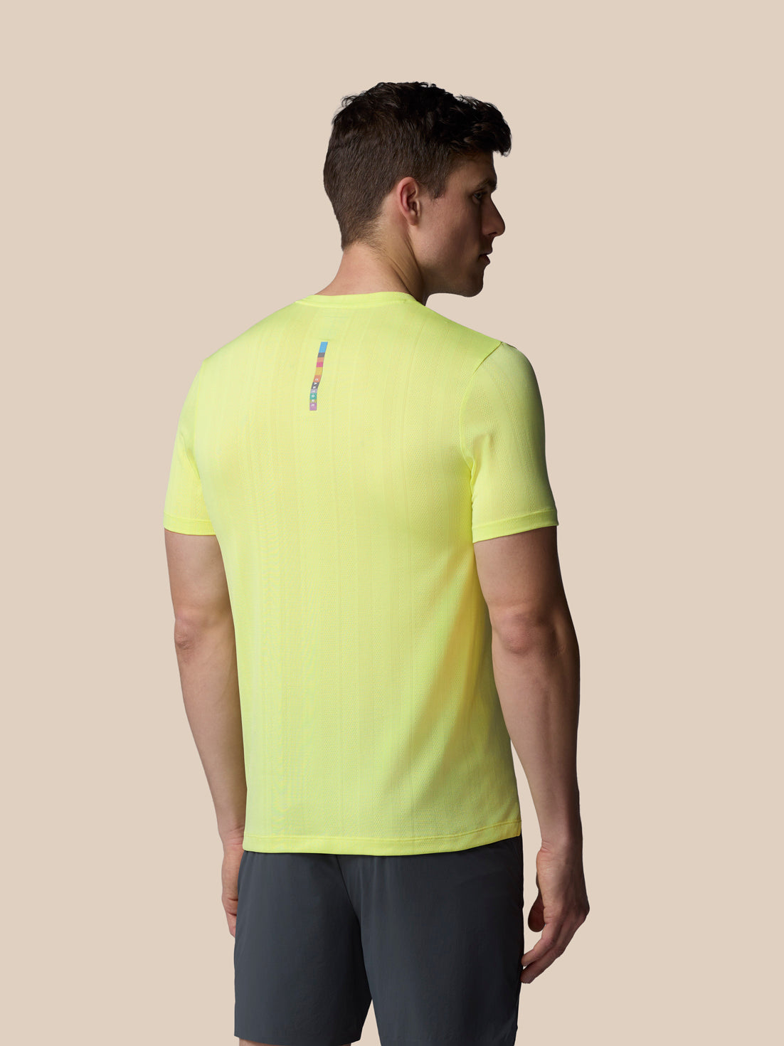 Man wearing a bright yellow t-shirt with a small logo on the back against a beige background