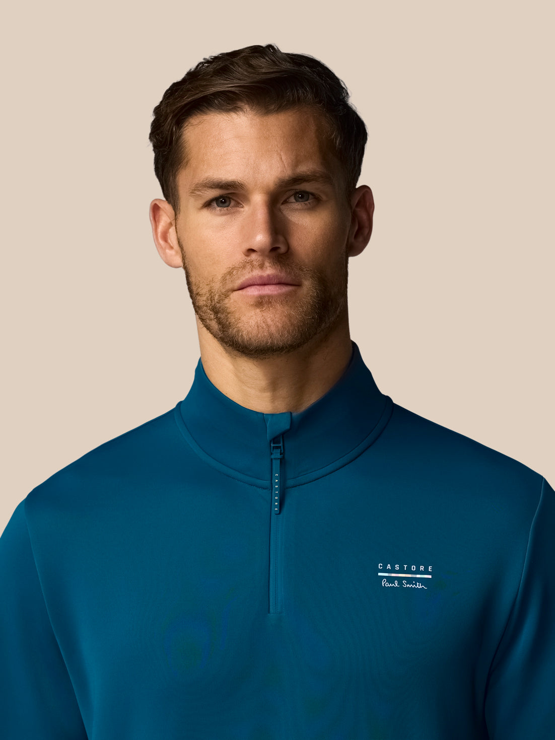 Man wearing a blue quarter-zip sweatshirt with a brand logo on a beige background