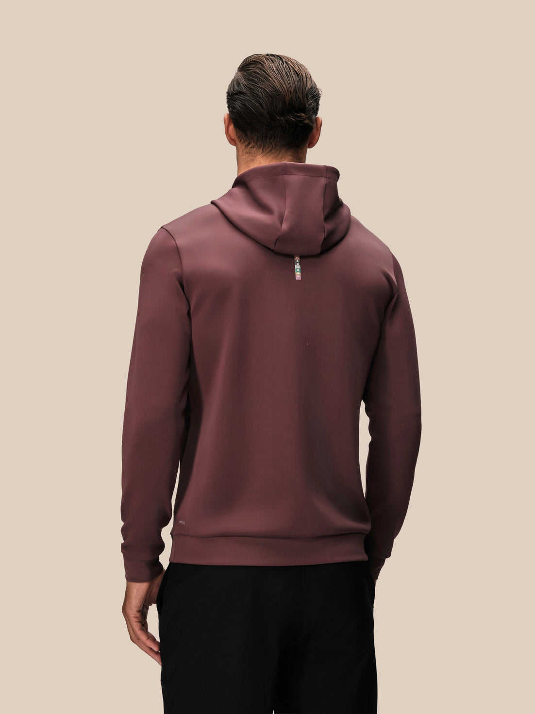 A man viewed from behind wearing a dark plum-brown hooded sweatshirt and black pants, standing against a solid beige background.