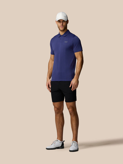 Man wearing a blue polo shirt, black shorts, and white shoes on a beige background