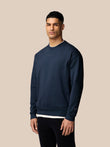 Male model wearing navy Adapt Lifestyle Sweatshirt with crew neck and oversized silhouette; long sleeves with ribbed cuffs and hem and a small chest logo with Castore wings on the left arm