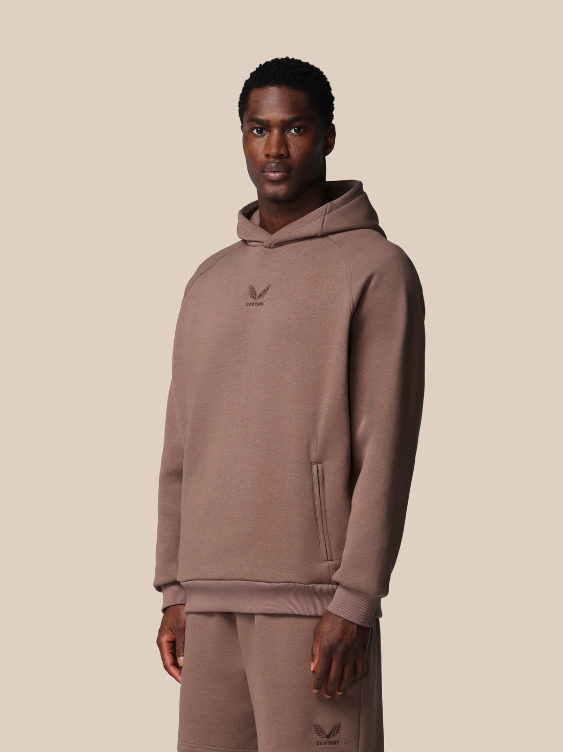Man wearing a brown hoodie and pants with a logo on a beige background