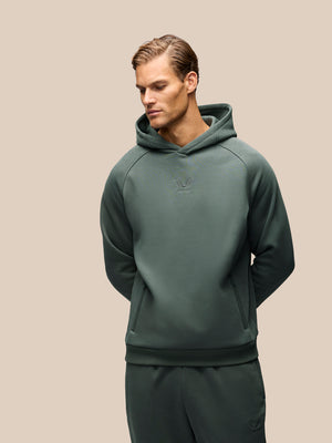 A man wearing a dark green pullover hoodie and matching sweatpants, photographed against a solid beige studio background.