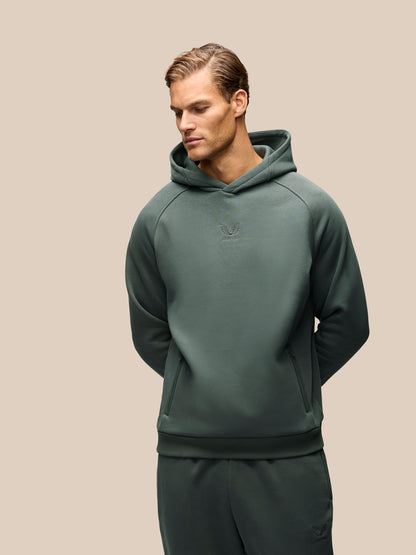 A man wearing a dark green pullover hoodie and matching sweatpants, photographed against a solid beige studio background.