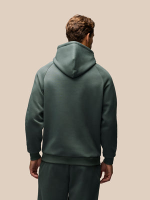 A man viewed from behind wearing a dark green hooded sweatshirt and matching sweatpants against a solid beige background.