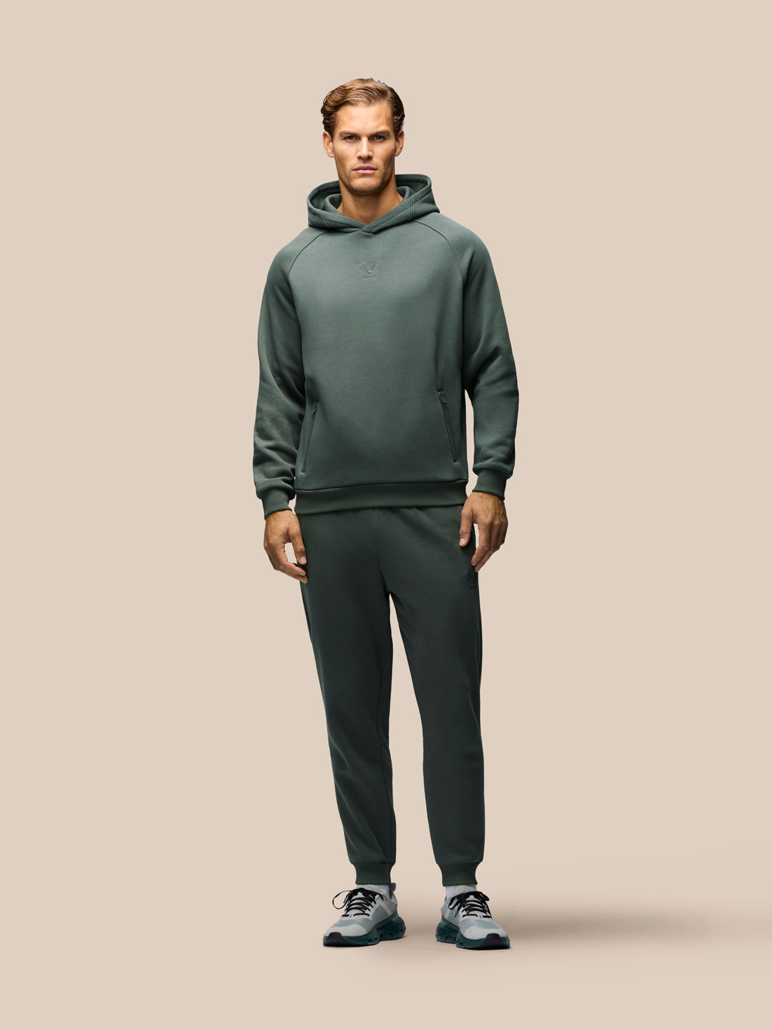 A man modeling a dark green athletic hoodie and matching jogger pants set, paired with gray and blue sneakers, against a solid beige background.