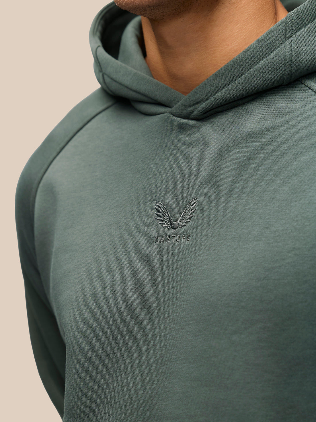 Close-up of a person wearing a muted sage green pullover hoodie with a tonal embroidered winged logo and the word 'CASTORE' on the chest, against a plain light beige background.