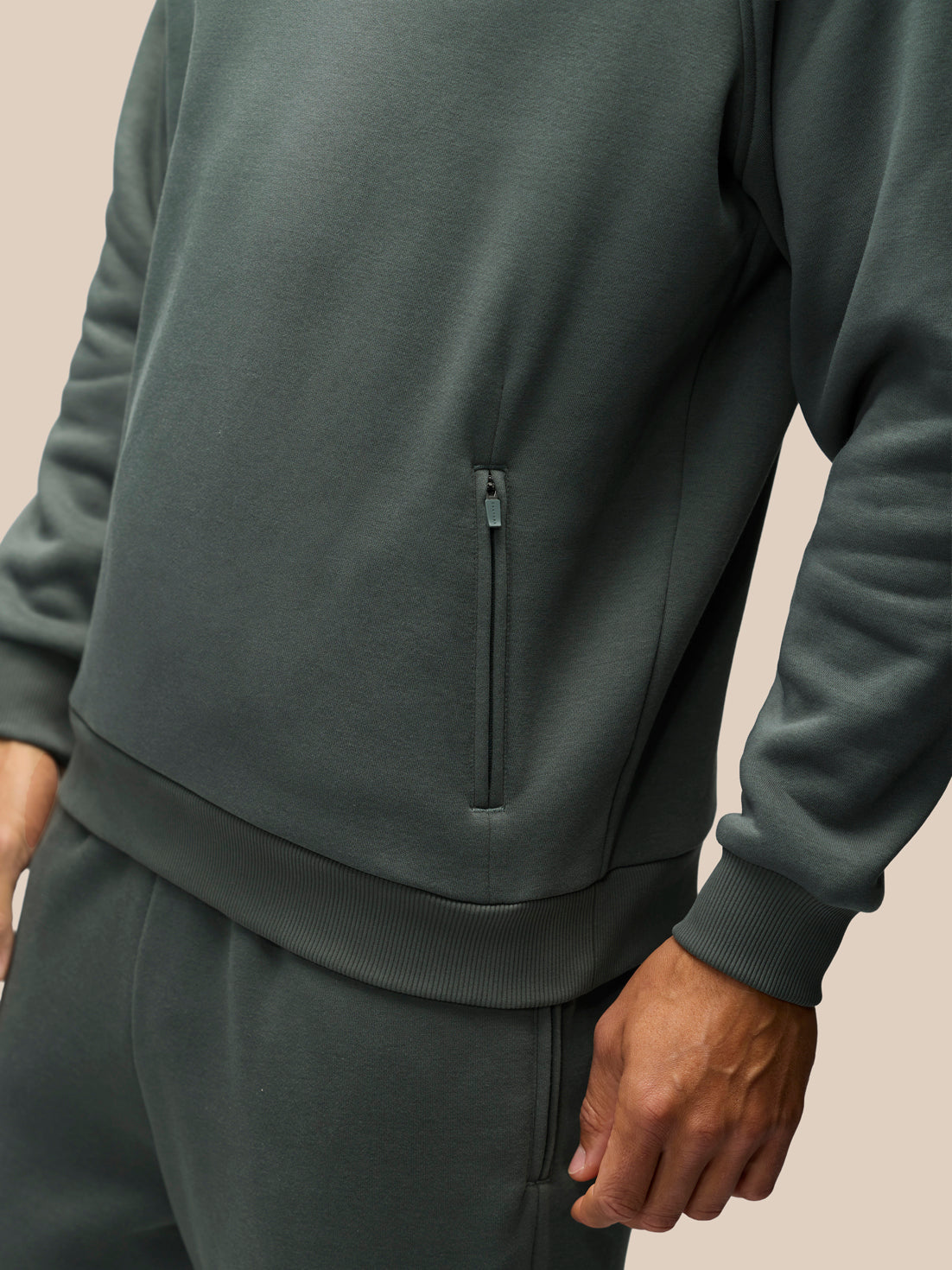 Close-up of a person wearing a dark gray-green zippered sweatshirt and matching sweatpants against a plain beige background.