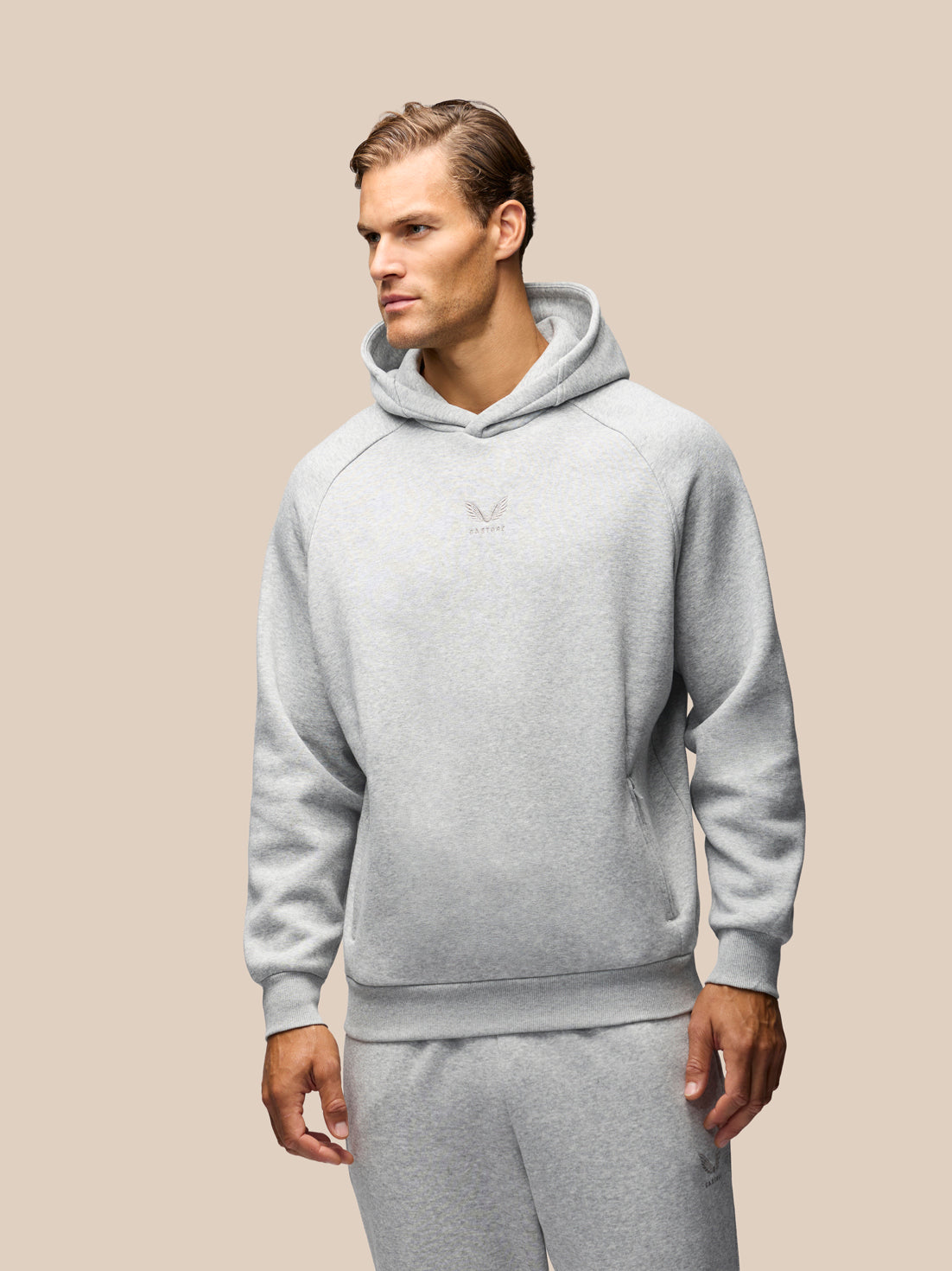 A man wearing a light gray heathered pullover hoodie and matching sweatpants, shown from the waist up against a solid beige background.