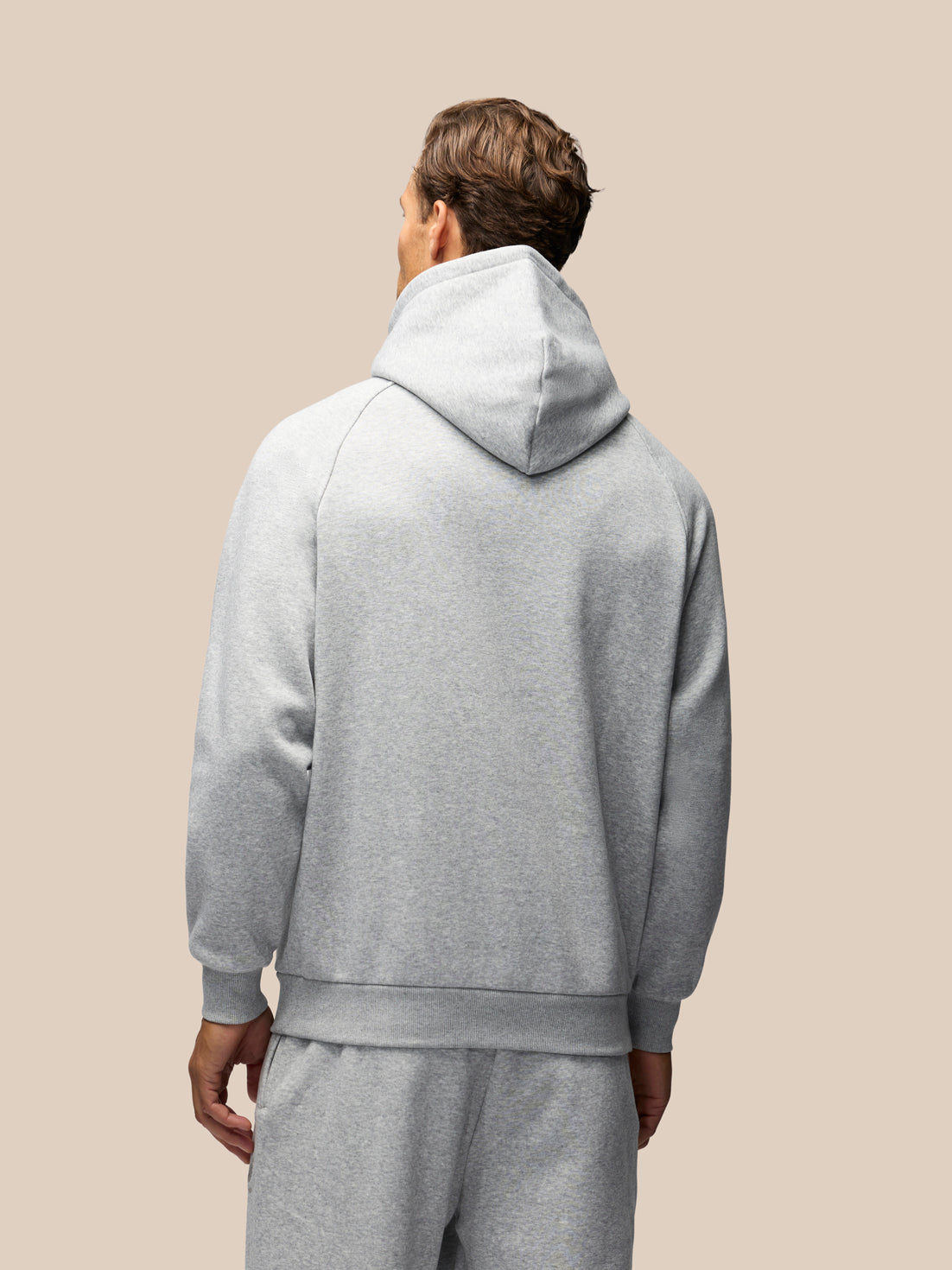 A man viewed from behind wearing a light gray heathered hooded sweatshirt and matching sweatpants against a solid beige background.
